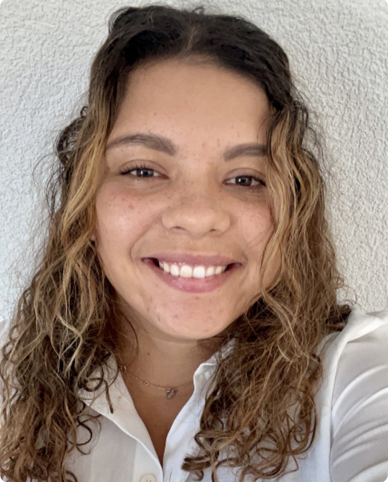 Jeune femme souriante avec des cheveux bouclés, portant une chemise blanche, devant un mur blanc.