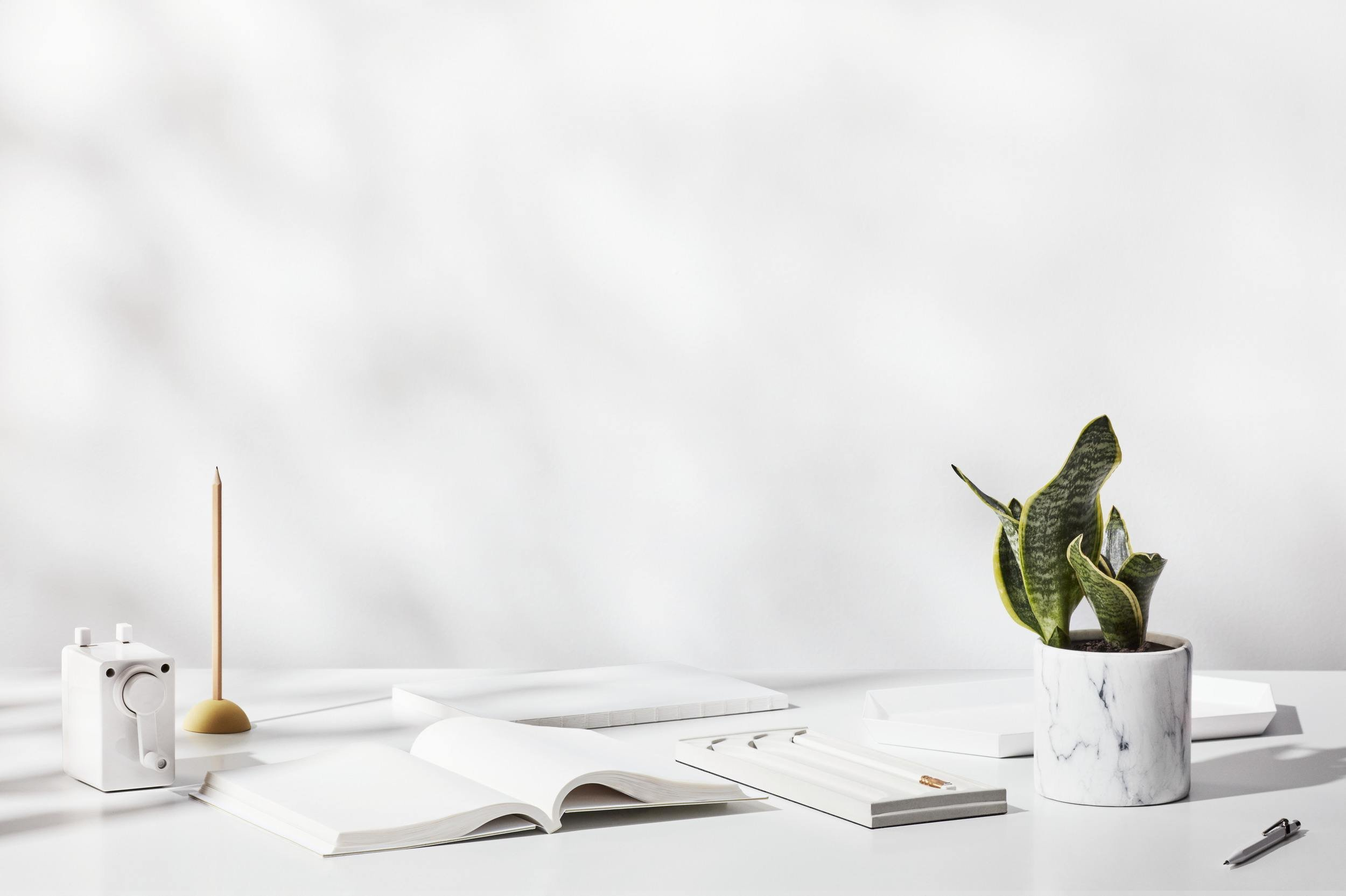 Bureau minimaliste blanc avec une plante en pot en marbre, un carnet ouvert, un stylo, un appareil photo blanc, une bougie, un bloc-notes et des papers.